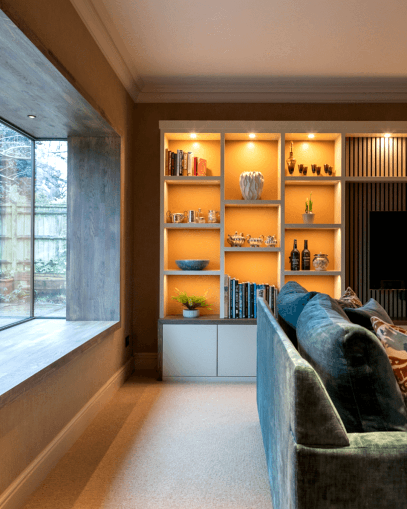 Contemporary living room with illuminated built-in shelving, modern sofa, warm neutral tones, and large picture window.