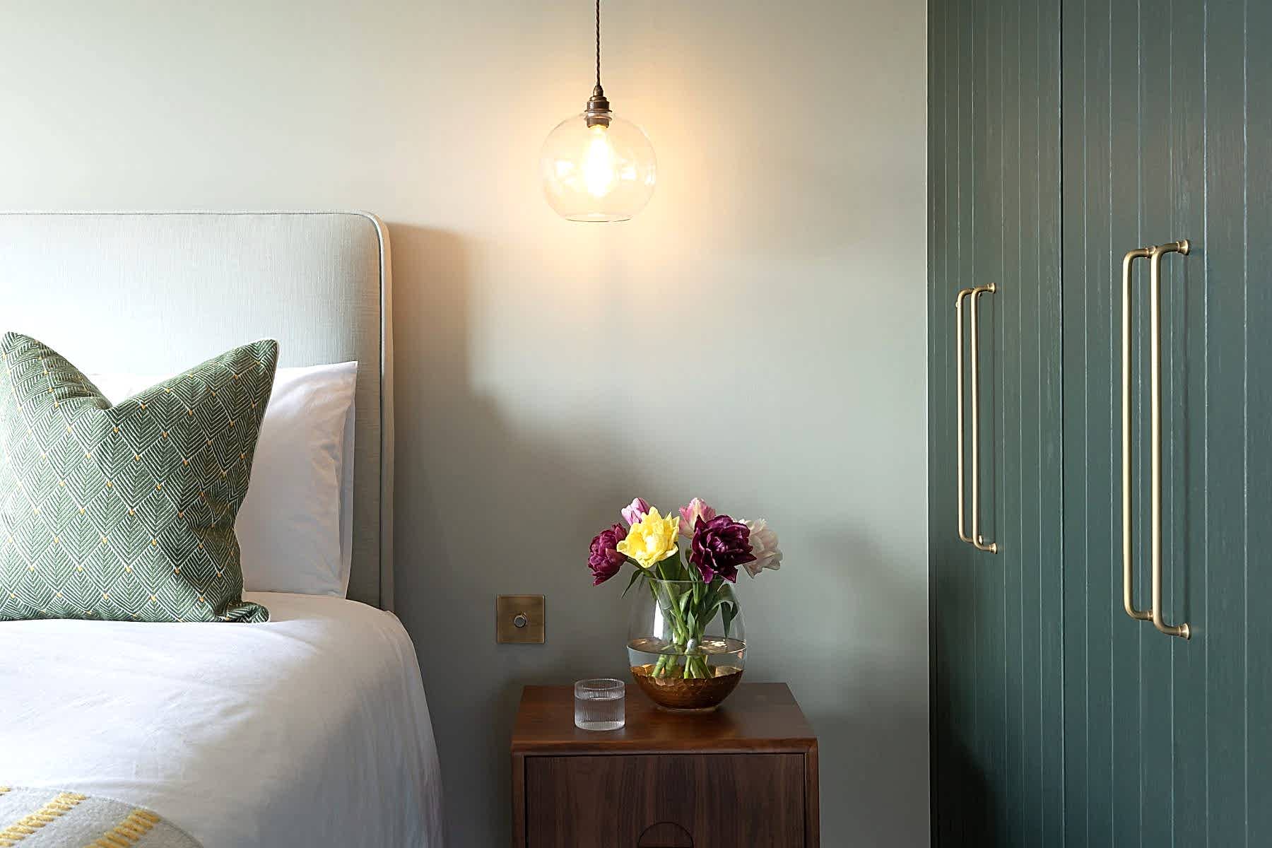 Minimalist bedroom corner with wooden bedside table, pendant light, and upholstered bed against a muted green wall.