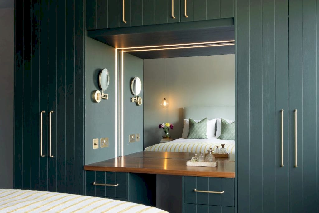 Dark green fitted wardrobes with integrated dressing table, illuminated mirror, and brass handles in a contemporary bedroom.