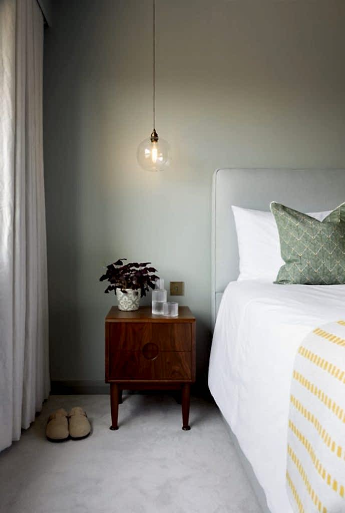 Minimalist bedroom corner with wooden bedside table, pendant light, and upholstered bed against a muted green wall.