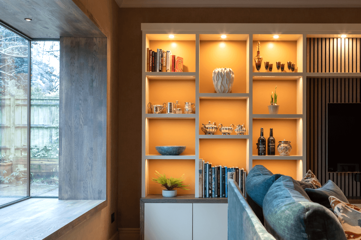 Contemporary living room with illuminated built-in shelving, modern sofa, warm neutral tones, and large picture window.