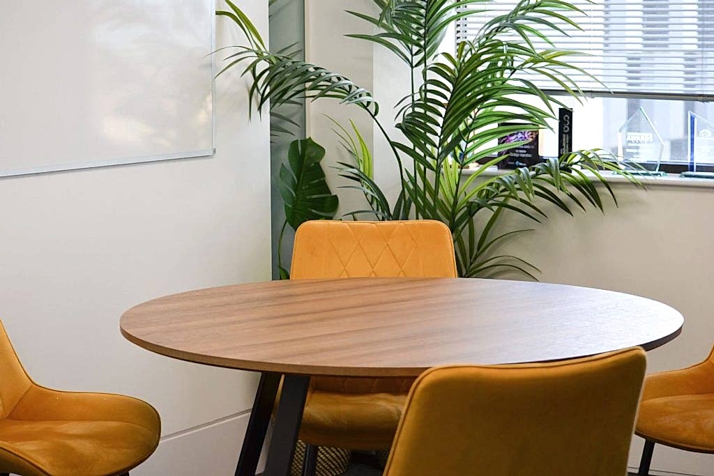 Round timber meeting table with mustard upholstered chairs and indoor plants in contemporary workspace.