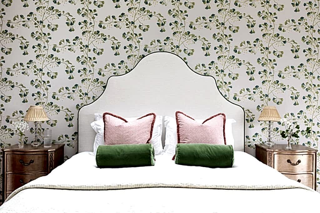 Bedroom with upholstered headboard, floral wallpaper, wooden bedside tables and layered cushions in soft pink and green tones.